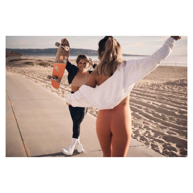 Two women walking on beach with skateboard. One woman faces forward in orange leggings. Other holds skateboard smiling.