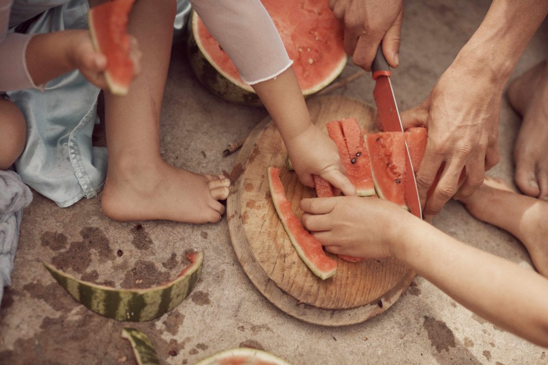 Overhead view of a group of children and an adult cutting and eating watermelon on a weathered concrete surface, close-up of hands and feet.