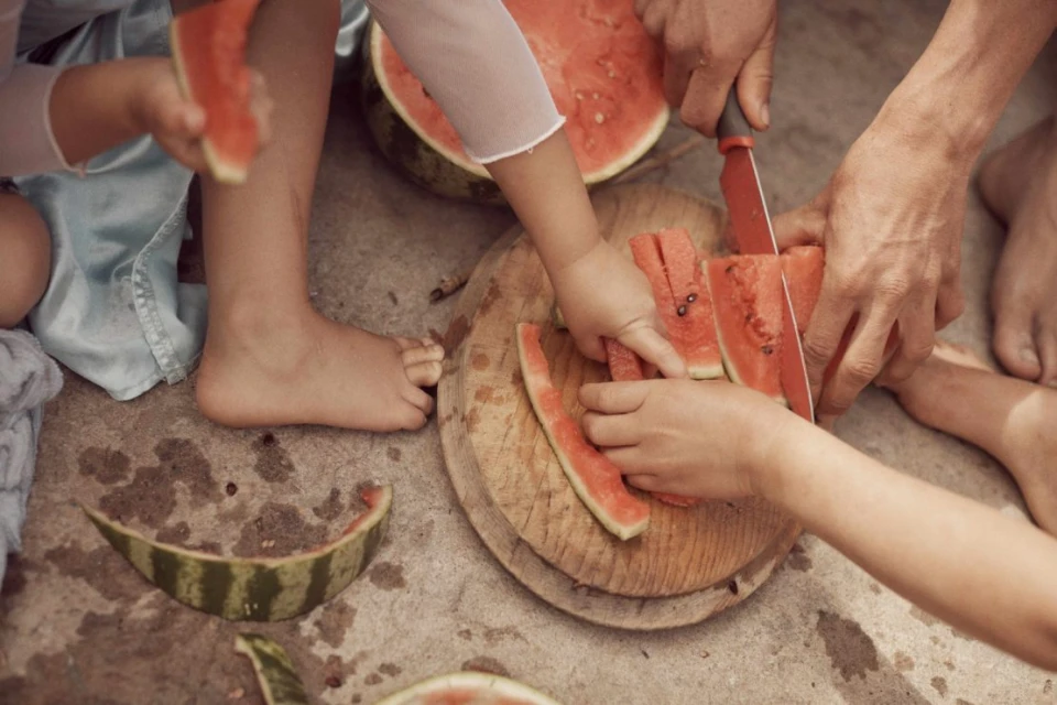 Overhead view of a group of children and an adult cutting and eating watermelon on a weathered concrete surface, close-up of hands and feet.