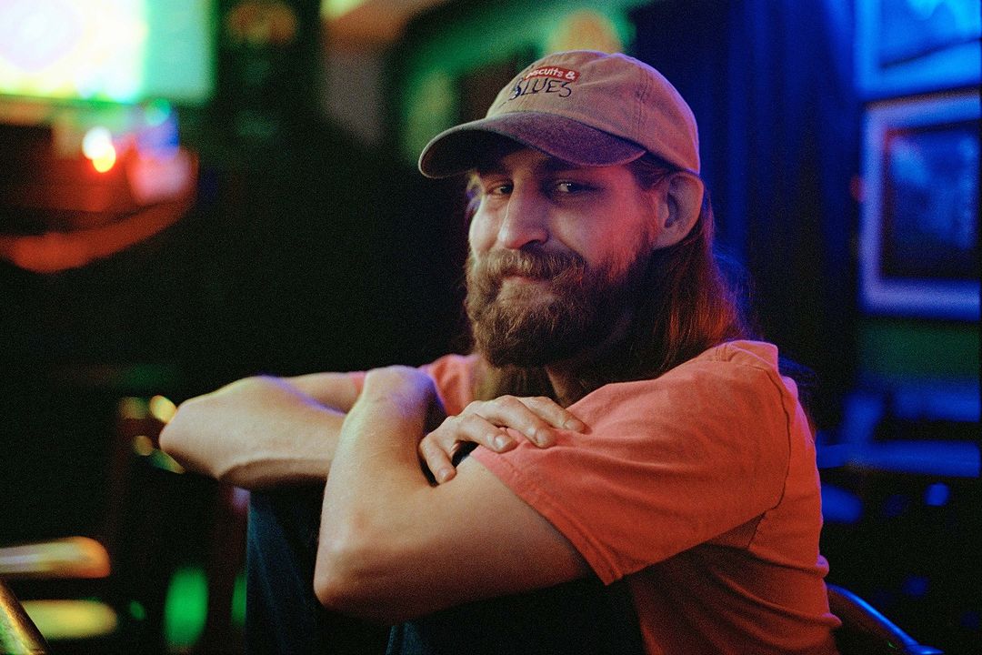 Adult man in a baseball cap with a beard sits indoors under blue and green neon lights, wearing a coral T-shirt and looking at the camera.