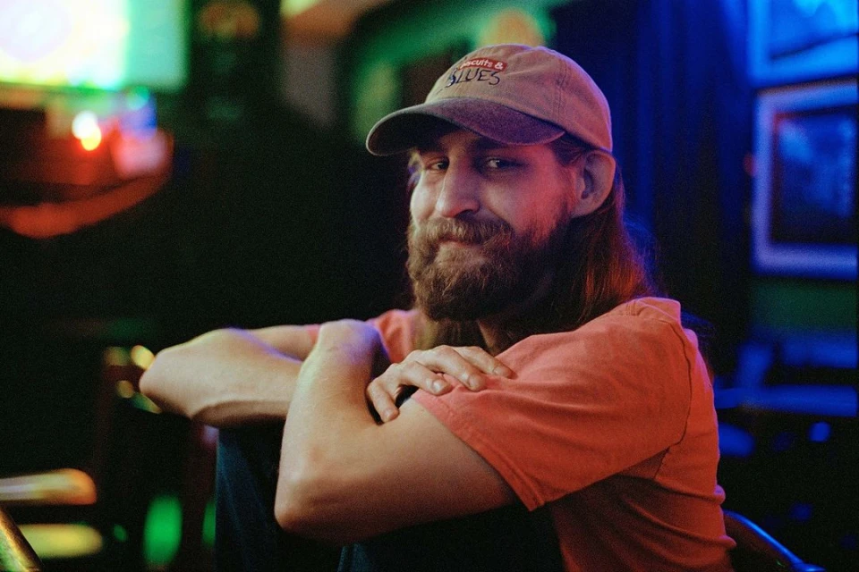 Adult man in a baseball cap with a beard sits indoors under blue and green neon lights, wearing a coral T-shirt and looking at the camera.