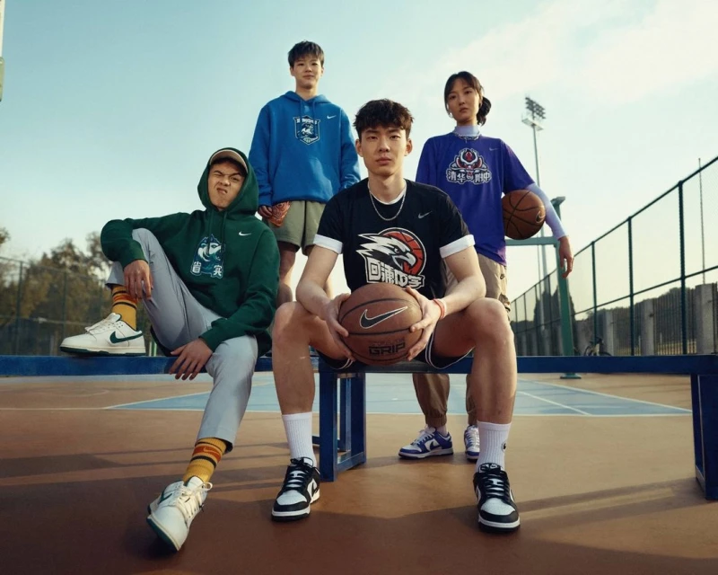 Four young people in athletic wear pose outdoors on a basketball court, displaying Nike branded apparel and holding a basketball.