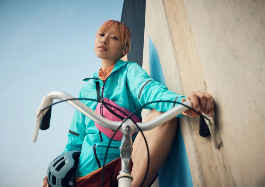 Woman with peach hair and bright windbreaker poses with a bicycle, helmet, and pink belt bag against a neutral architectural background.