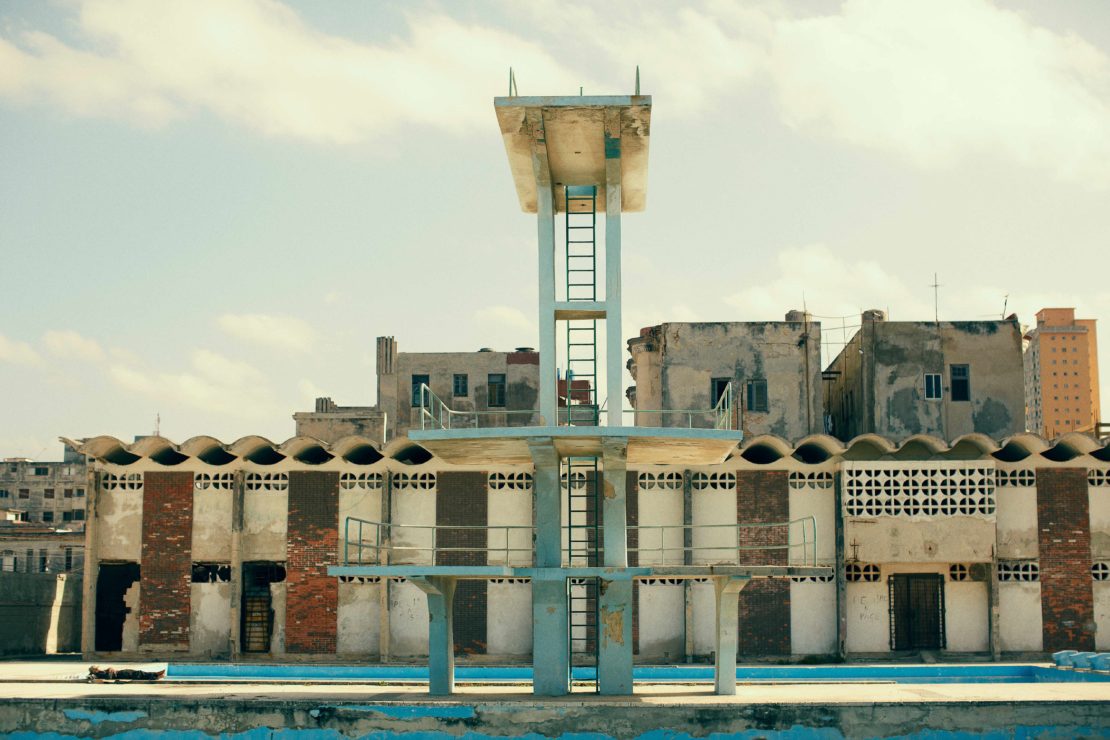Swimming pool diving platform under a cloudy sky in an urban environment; derelict condition with peeling paint and aged infrastructure.