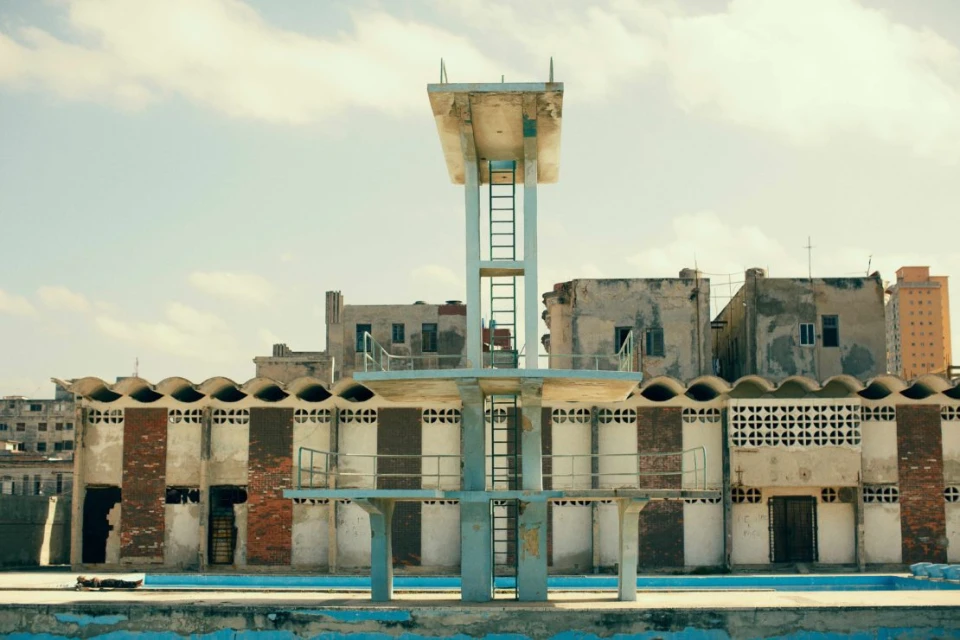 Swimming pool diving platform under a cloudy sky in an urban environment; derelict condition with peeling paint and aged infrastructure.