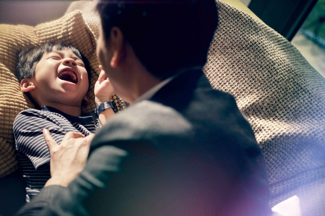 Father and son laughing indoors, the child lying on a cushion and father standing nearby, wearing a suit jacket, natural light.