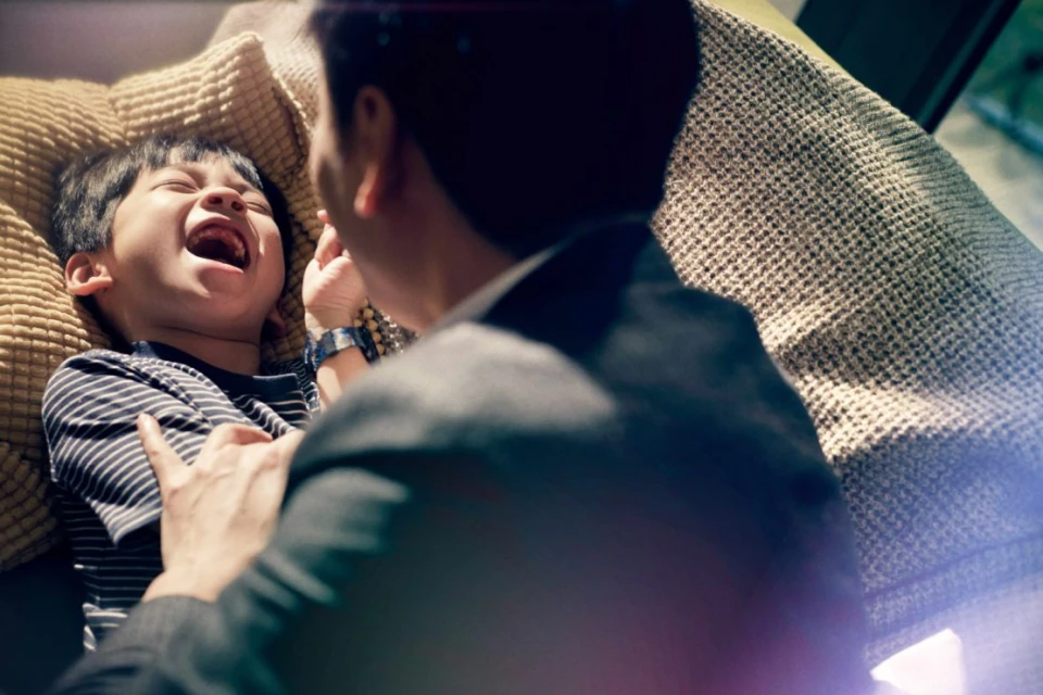 Father and son laughing indoors, the child lying on a cushion and father standing nearby, wearing a suit jacket, natural light.