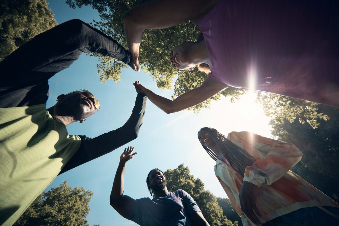 Diverse group of four people in circle, reaching up high-fiving against a blue sky and green trees, low angle, positive mood, teamwork.