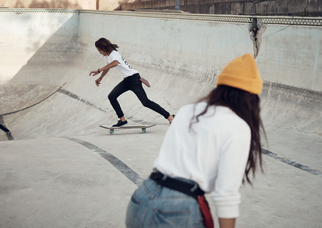Young adult skateboarding in a concrete skatepark, photographed at eye-level with a second person in the foreground wearing a yellow hat.