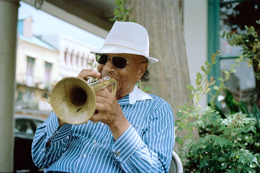 An adult male plays a brass trumpet while seated outdoors. He wears a white hat and sunglasses, with a blurred building in background.