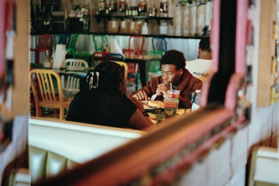 Two people sit at a restaurant table. An African-American man eats while a woman sits opposite him. Casual indoor dining with colorful chairs.