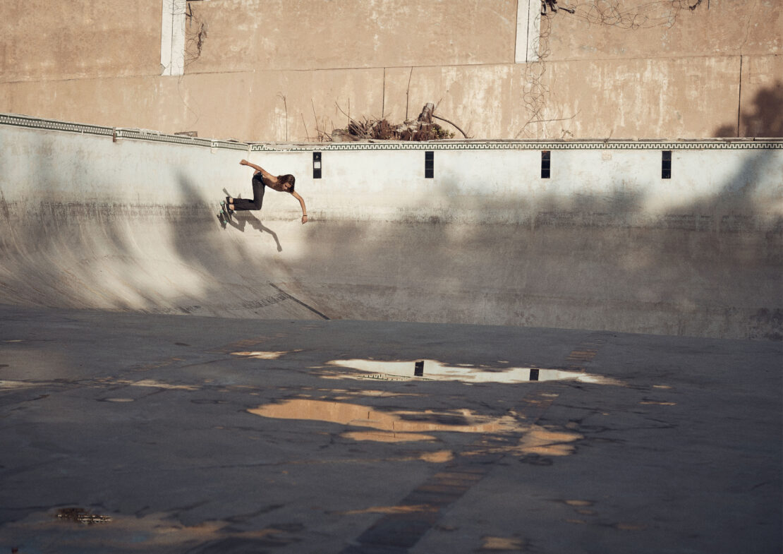 A skateboarder performs a trick in an empty concrete pool under bright sunlight, casting a long shadow, creating a dynamic action shot.