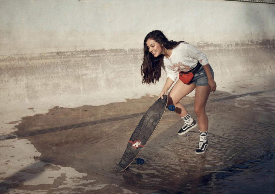 Young woman with long dark hair poses with a skateboard in a concrete pool, wearing jean shorts and a white long-sleeve shirt.