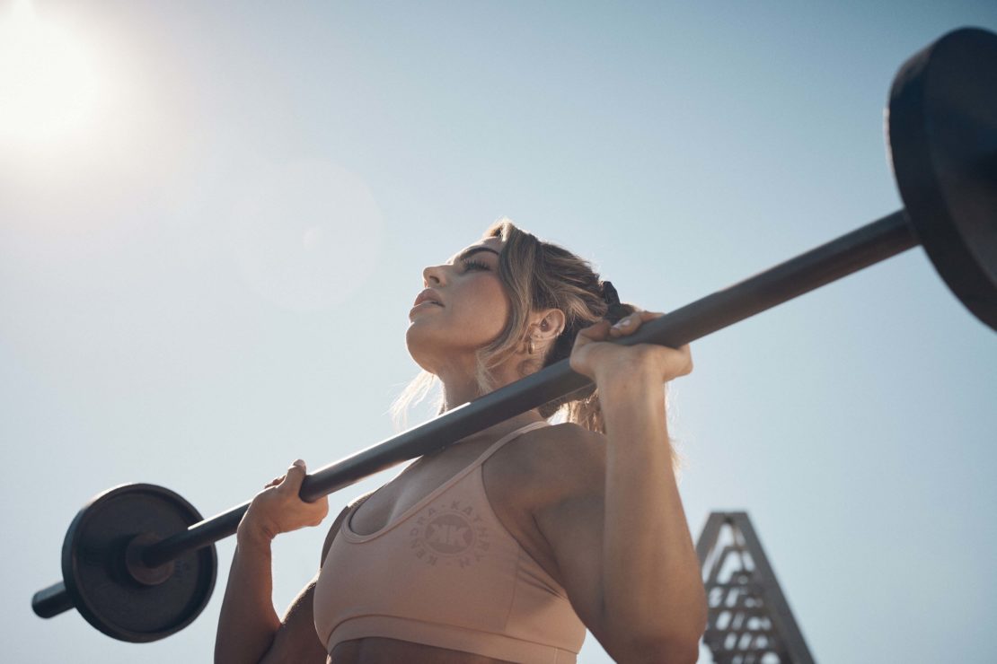 Adult female lifting barbell overhead in outdoor fitness shot with blue sky. Woman in sportswear exercising with weights on sunny day.