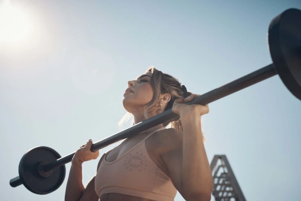 Adult female lifting barbell overhead in outdoor fitness shot with blue sky. Woman in sportswear exercising with weights on sunny day.
