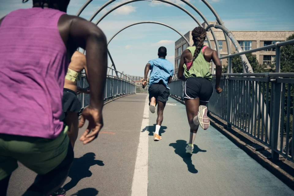 Group of diverse adult runners in athletic wear training outdoors on a grey pedestrian bridge with an arched metal structure overhead.