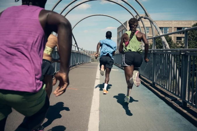 Group of diverse adult runners in athletic wear training outdoors on a grey pedestrian bridge with an arched metal structure overhead.