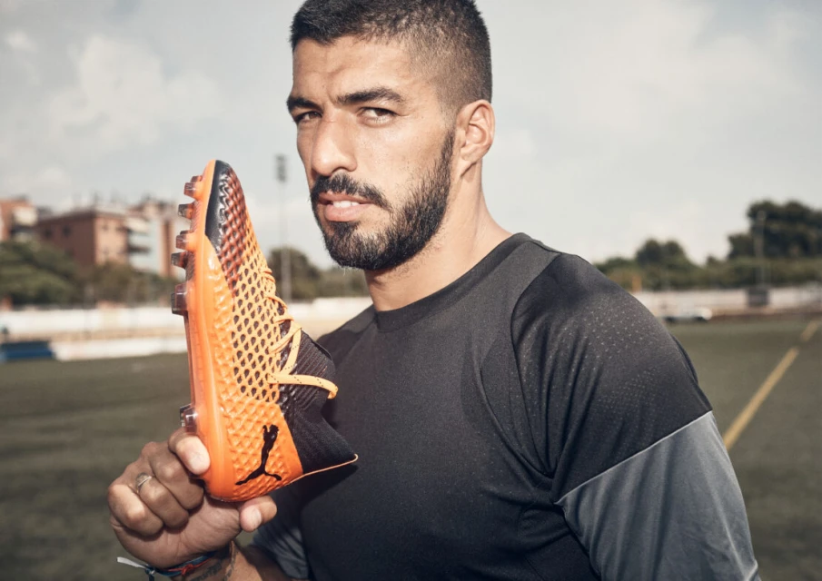 Close-up of soccer player Luis Suarez holding a Puma soccer cleat on a grassy field with buildings in the background under a cloudy sky.