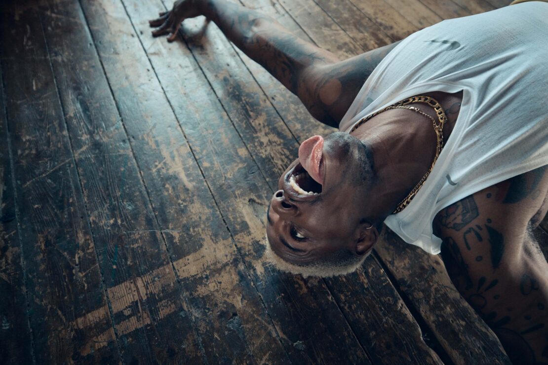 Person with bleached hair lying on their back on a distressed wood floor with their mouth open and tongue sticking out.