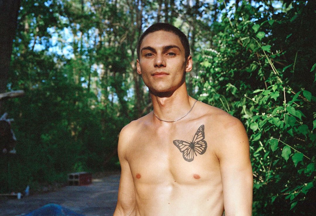 Adult male poses shirtless outdoors with a butterfly tattoo on his chest, set against a leafy green background in natural light.