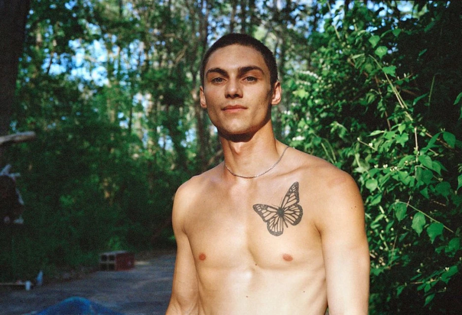 Adult male poses shirtless outdoors with a butterfly tattoo on his chest, set against a leafy green background in natural light.