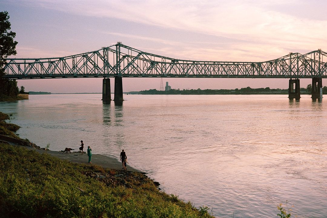 Truss bridge spans wide river as people gather on the grassy shoreline at dusk, with calm water reflecting the fading light.