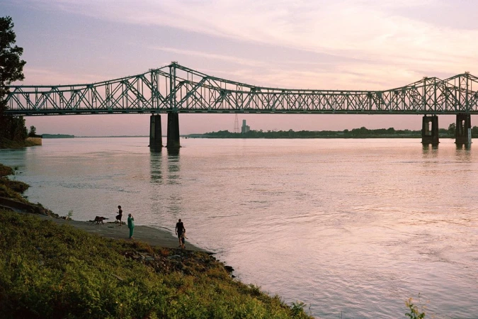 Truss bridge spans wide river as people gather on the grassy shoreline at dusk, with calm water reflecting the fading light.
