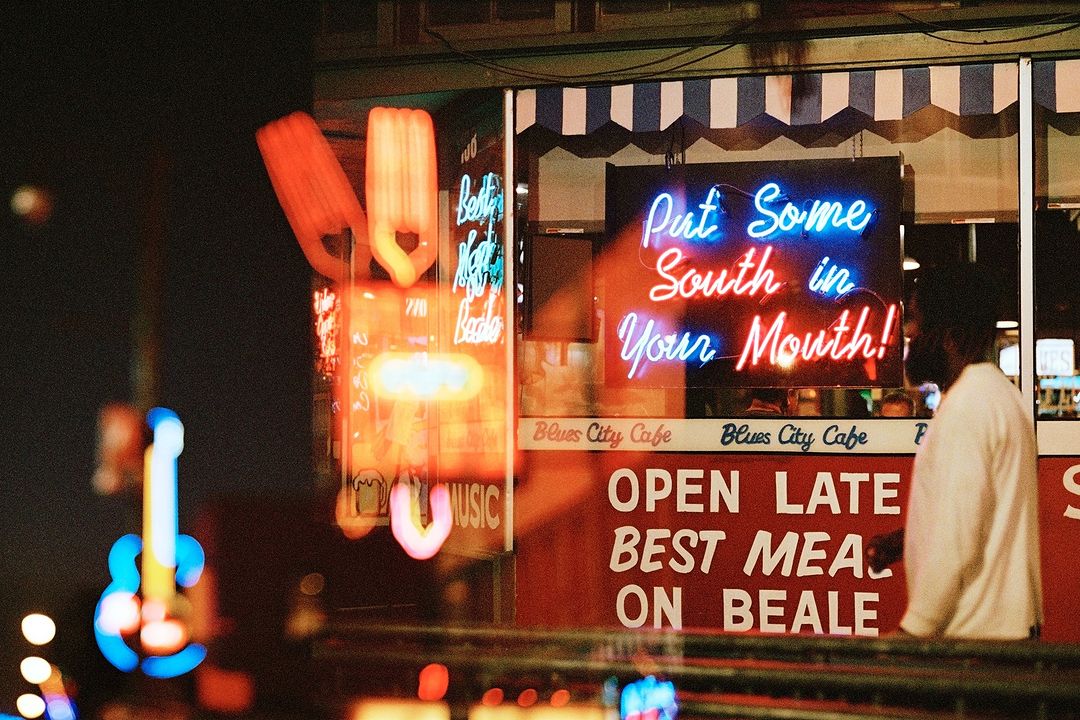 Neon signs illuminate Blues City Cafe in Memphis at night. One sign promotes southern cuisine while a blurred man walks nearby.
