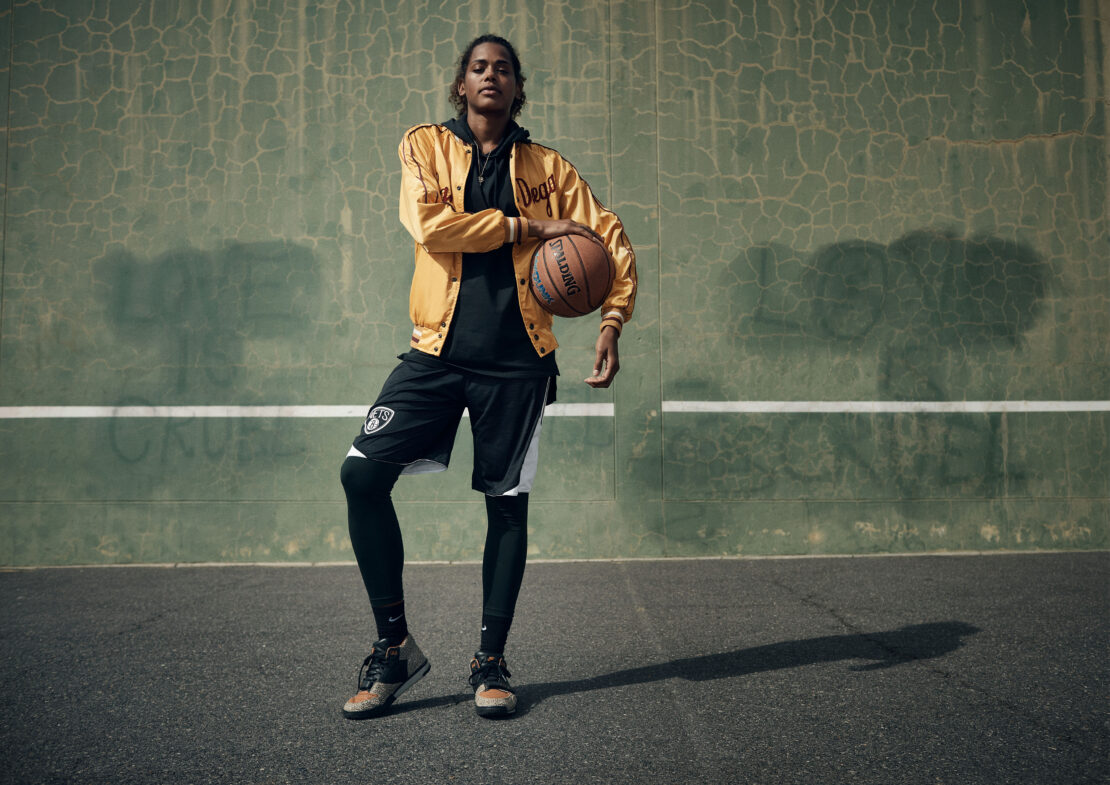 A female basketball player in athletic wear holds a basketball on an outdoor court against a textured green wall in the daytime.