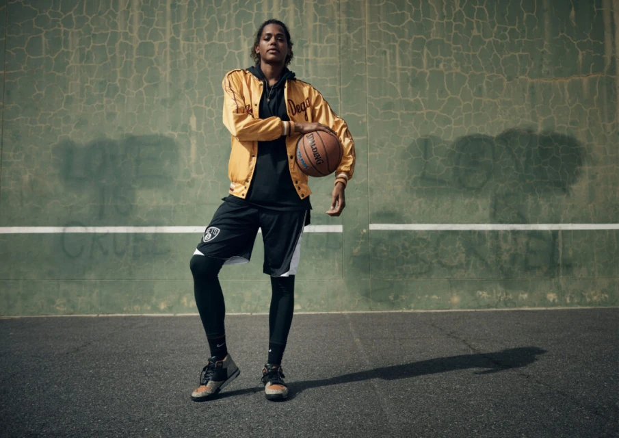 A female basketball player in athletic wear holds a basketball on an outdoor court against a textured green wall in the daytime.