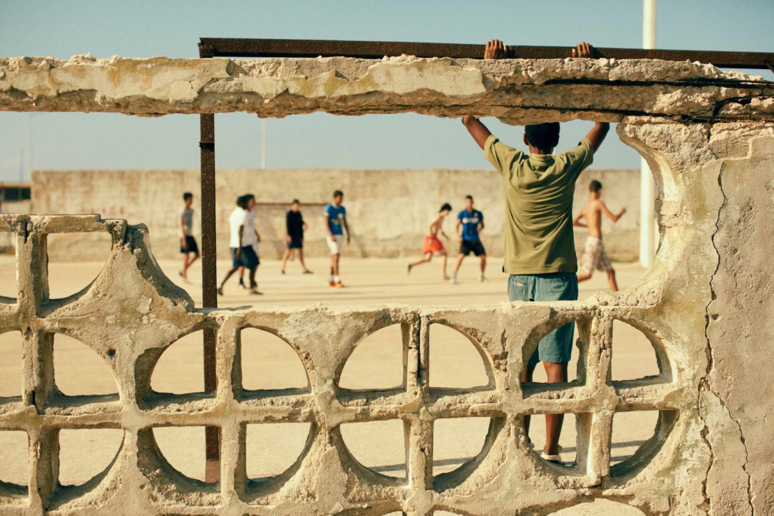 Boys play soccer on a sandy lot framed by a weathered wall. One boy supports himself on the crumbling structure, overlooking the game.