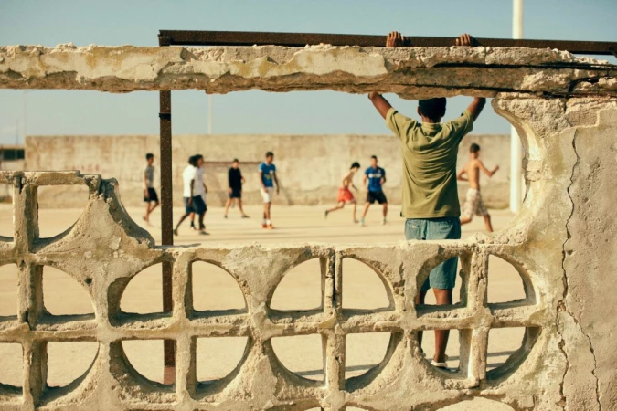 Boys play soccer on a sandy lot framed by a weathered wall. One boy supports himself on the crumbling structure, overlooking the game.