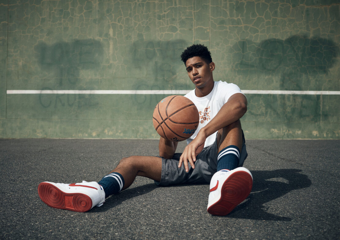 Young adult male poses with a basketball on an outdoor court. He's wearing a white tee, grey shorts, tube socks and athletic shoes.