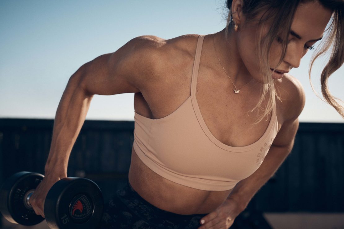 A woman in workout clothes lifts a dumbbell. The image captures the muscular definition in her arms and back in an outdoor setting.