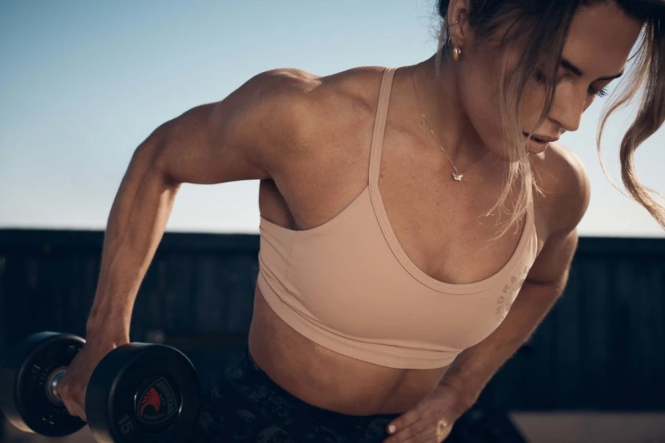 A woman in workout clothes lifts a dumbbell. The image captures the muscular definition in her arms and back in an outdoor setting.