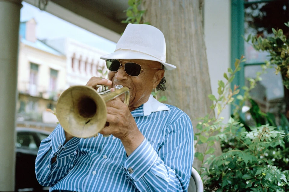 An elderly African-American man in sunglasses and a white hat plays a brass trumpet outdoors with a building and greenery in the background.