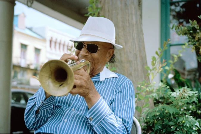 An elderly African-American man in sunglasses and a white hat plays a brass trumpet outdoors with a building and greenery in the background.