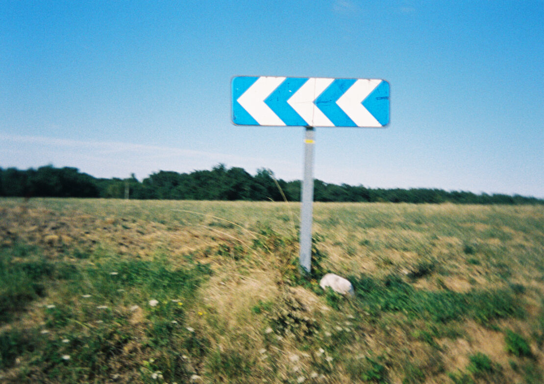 Daytime shot of blue and white directional road sign in rural setting with grassy field and line of trees under clear blue sky.