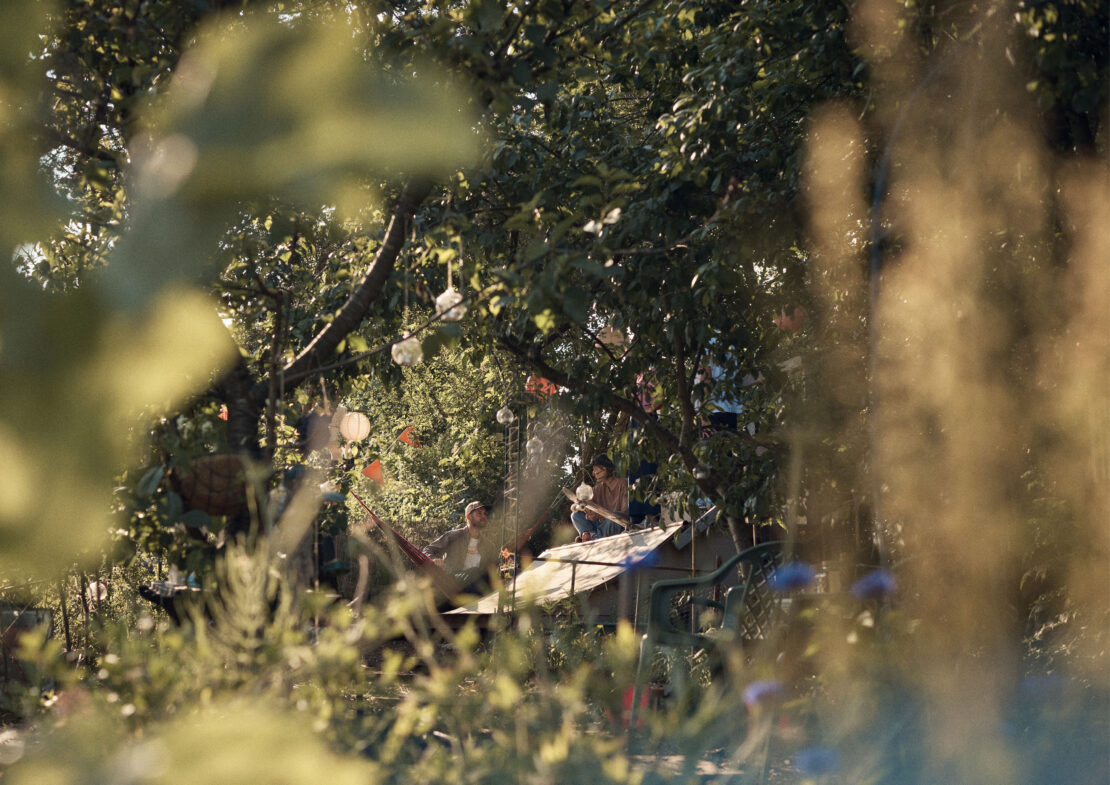 Two people sitting on a rooftop surrounded by lush green trees and foliage, viewed through foreground plants and flowers, in a sunny setting.