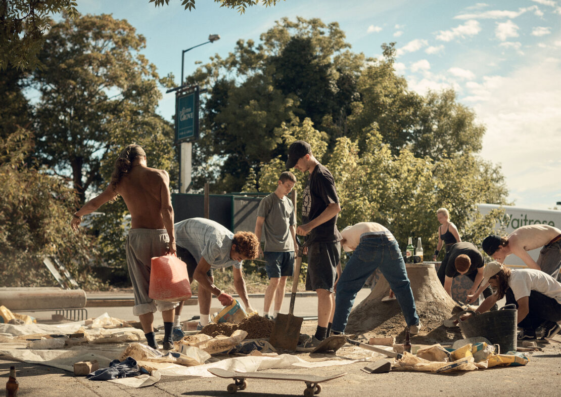 Group of young adults working on a concrete skateboarding ramp outdoors on a sunny day, trees and building in the background.