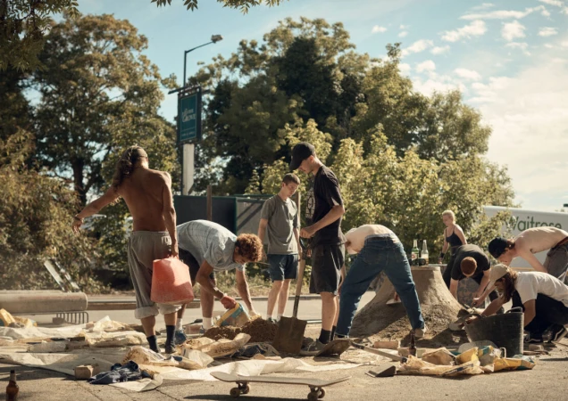 Group of young adults working on a concrete skateboarding ramp outdoors on a sunny day, trees and building in the background.