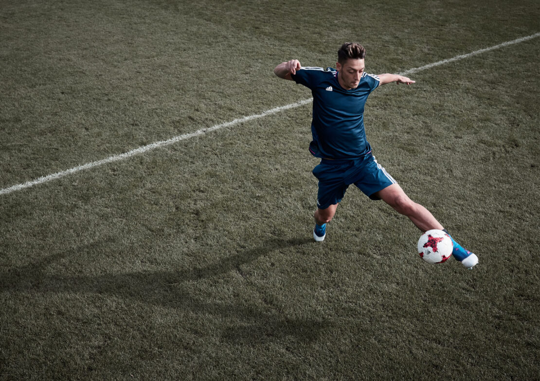 Male soccer player kicks ball on green field with white lines. He wears blue jersey, blue shorts, and blue and white shoes.