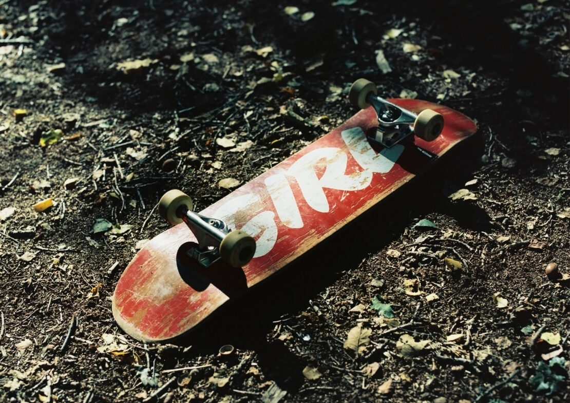 A red skateboard with white lettering lies on a forest floor scattered with twigs and leaves, capturing a grounded, urban sport scene.