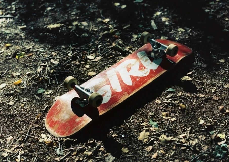 A red skateboard with white lettering lies on a forest floor scattered with twigs and leaves, capturing a grounded, urban sport scene.
