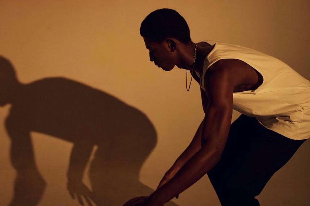 Athletic Black man in tank top prepares to dribble basketball in warm lighting, creating a dramatic shadow against a neutral wall.