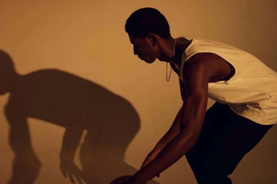 Athletic Black man in tank top prepares to dribble basketball in warm lighting, creating a dramatic shadow against a neutral wall.