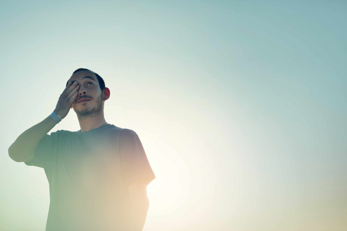 Man in grey t-shirt covering his eye against a bright sky background captured by Olly Burn