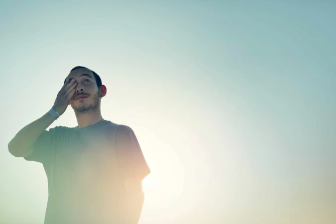 Man in grey t-shirt covering his eye against a bright sky background captured by Olly Burn