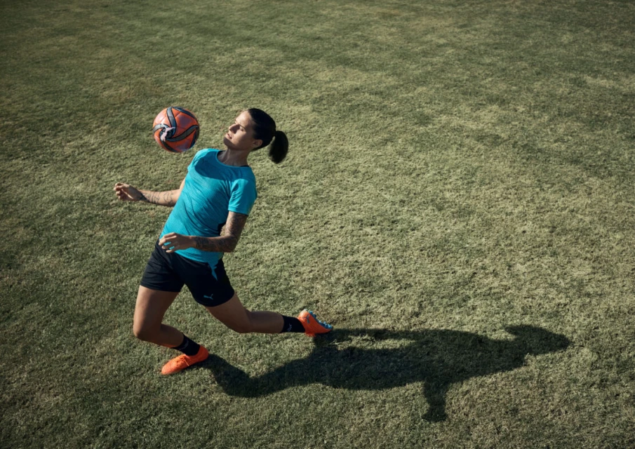 Woman in athletic wear balances a soccer ball on her head in a grassy field, captured from an overhead perspective on a sunny day.