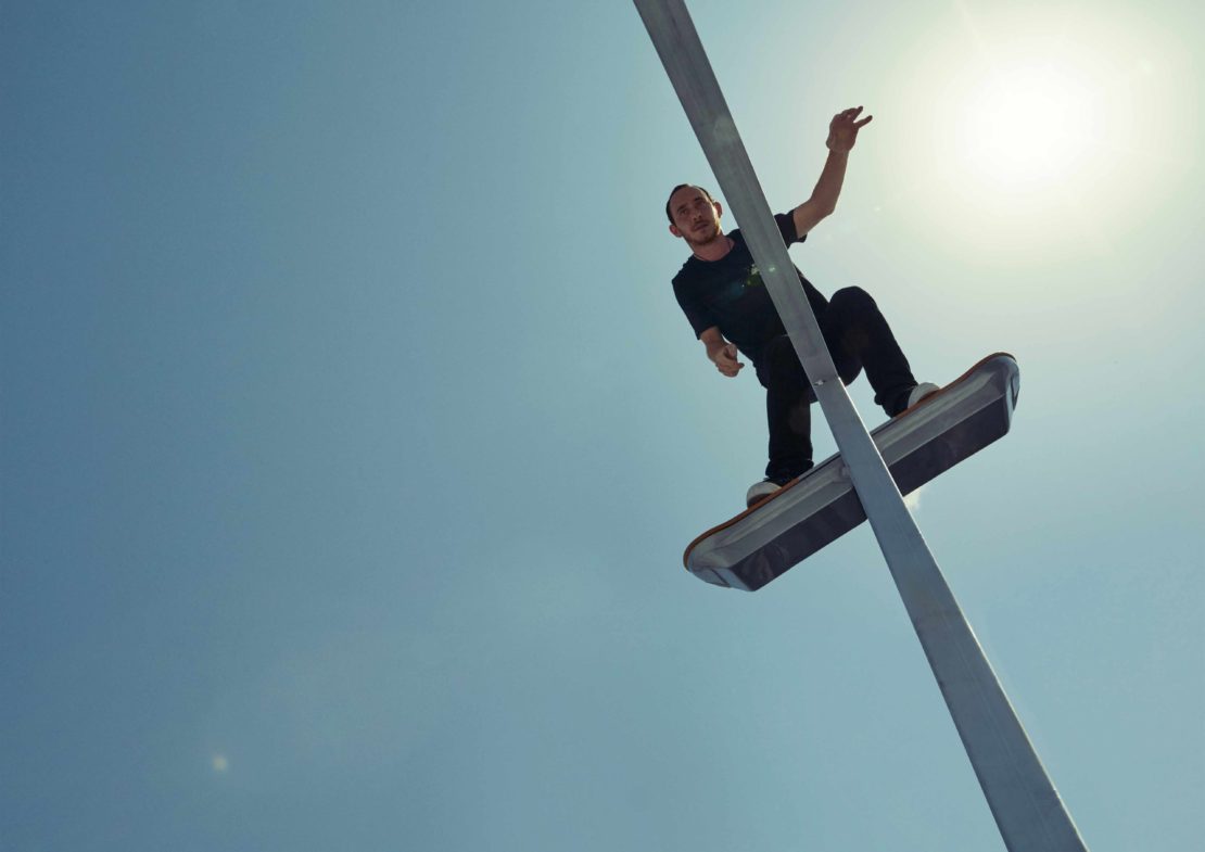 A low-angle shot captures a male skateboarder balancing on a street light, set against a clear blue sky with the sun shining brightly.